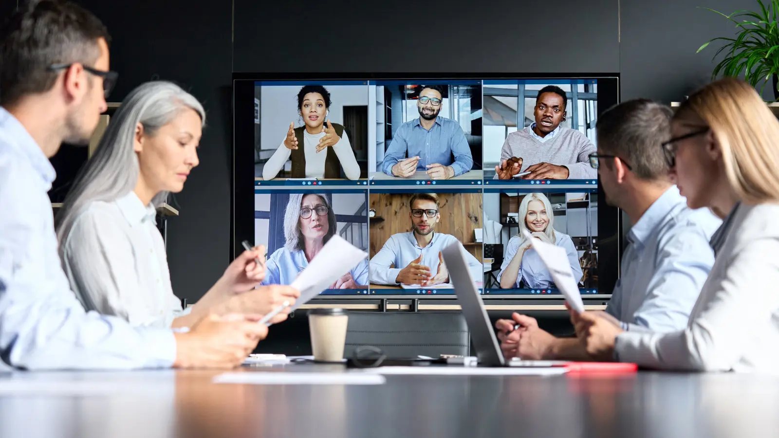 Photo of a board meeting with some people sitting around a table and some people on a Zoom screen.