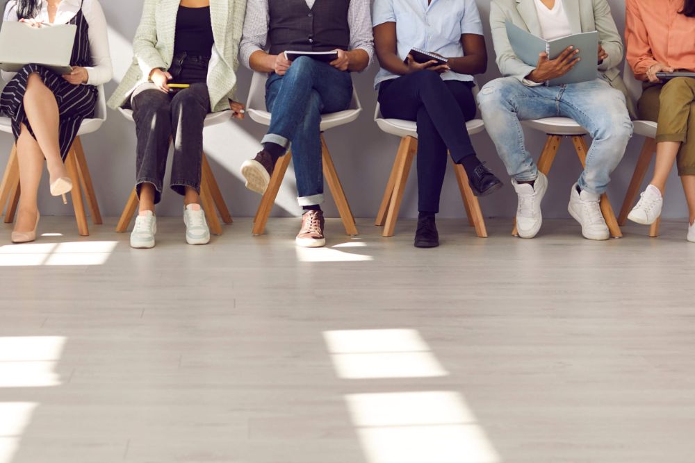 A line of job candidates sitting in chairs against a wall, just showing their legs.