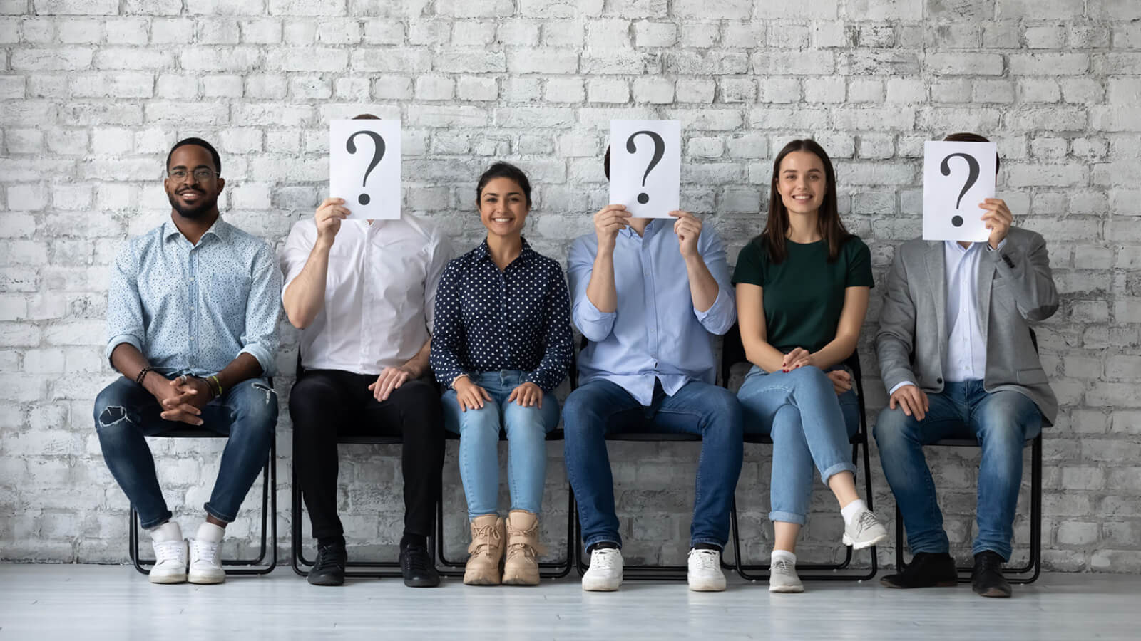A diverse group of job candidates sitting in chairs lined up against a brick wall. Some of them are holding question mark signs in front of their faces.