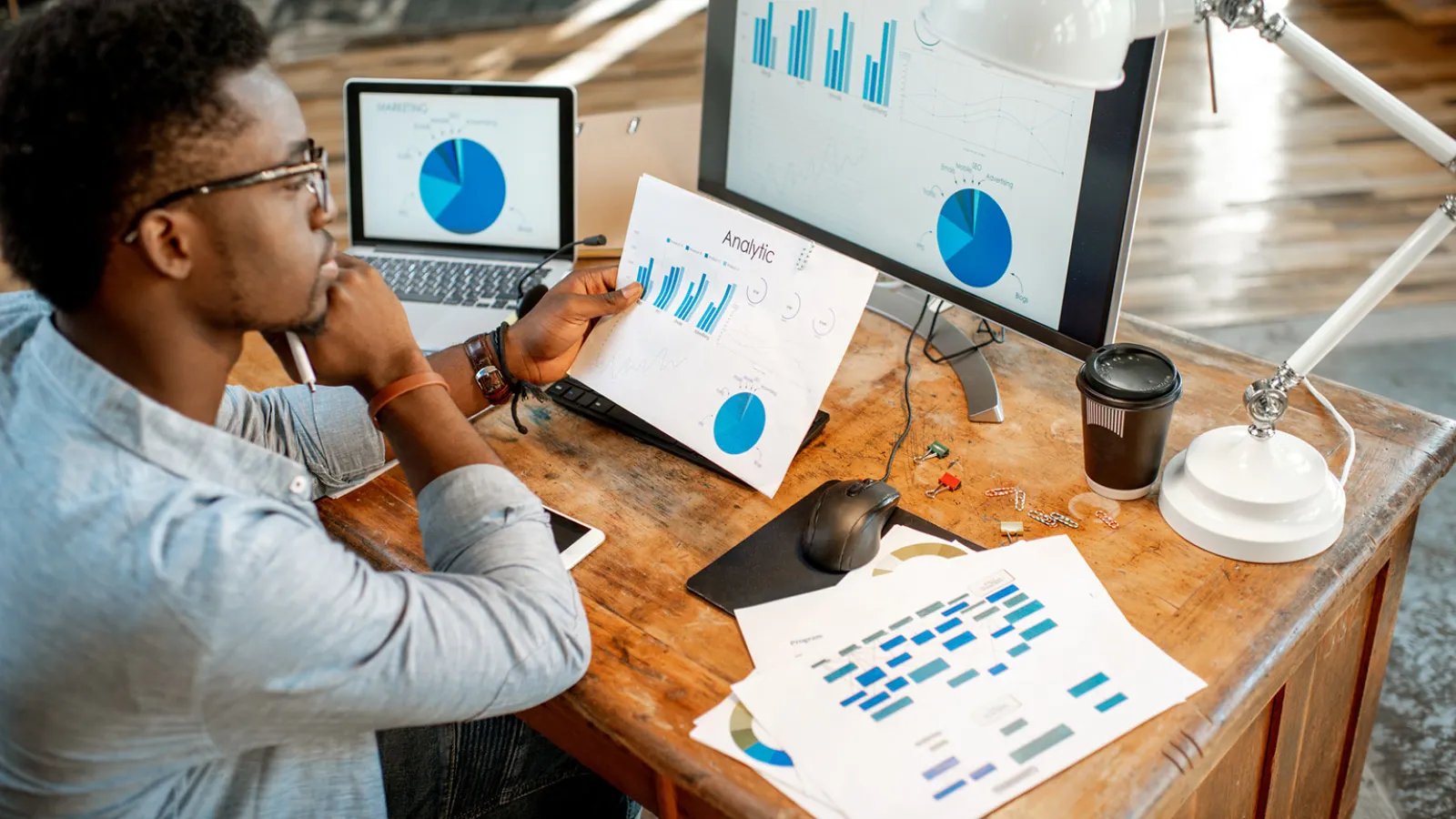 Man working at a computer holding samples of work with graphs and charts.