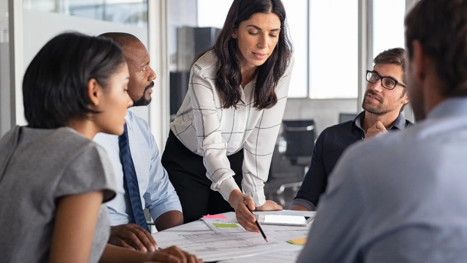 Photo of a work team sitting at a table with one person standing and pointing at some documents.