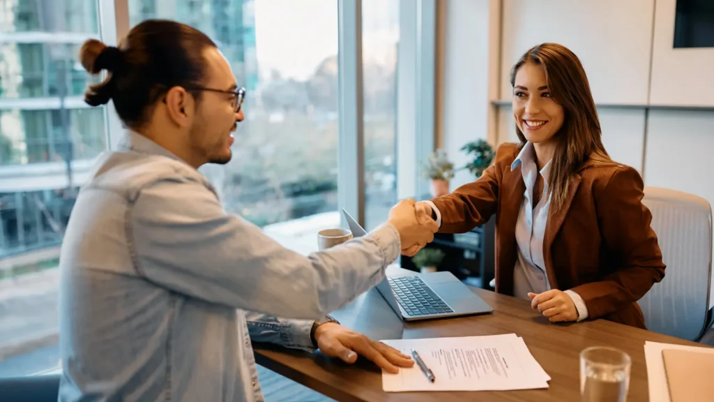 Photo of a female hiring manager shaking hands with a male job candidate. They are sitting across from each other at a desk.