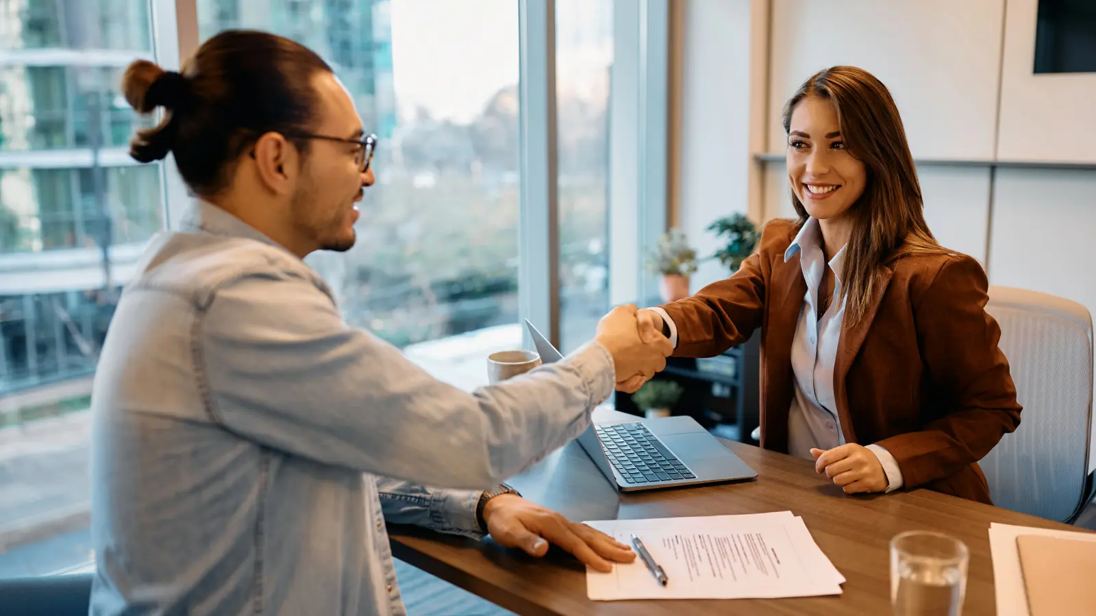 Photo of a female hiring manager shaking hands with a male job candidate. They are sitting across from each other at a desk.