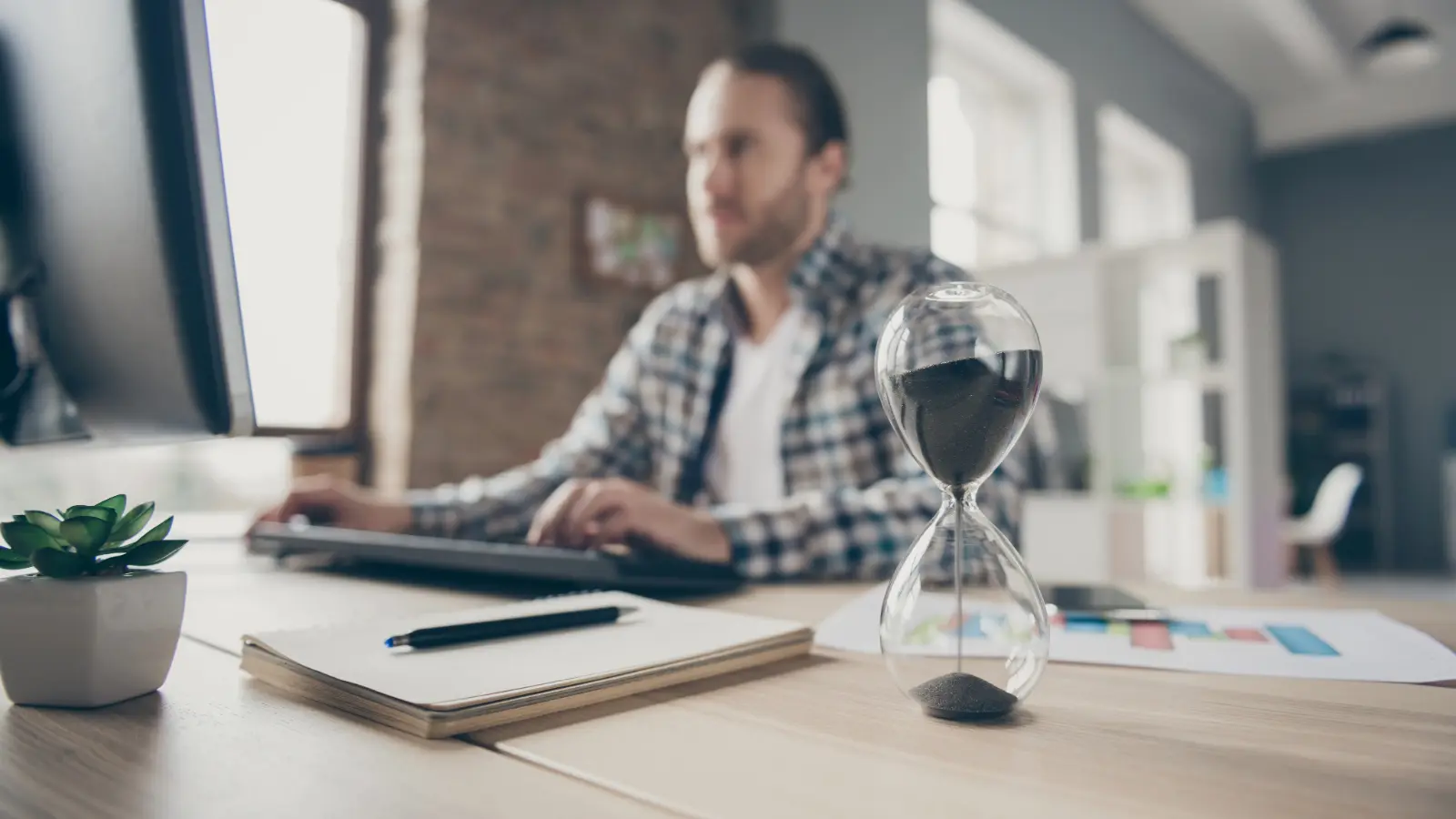 An hourglass in sharp focus showing time ticking down with a man working on a desktop computer in the background.