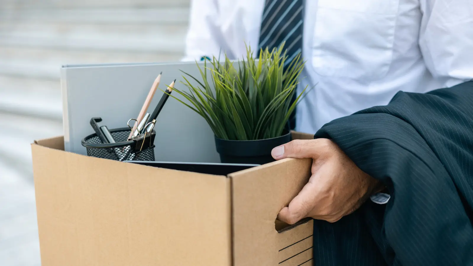 Close-up photo of a male executive's hands holding a box of personal items from his desk, showing that he has lost his job.