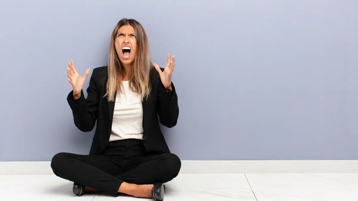 Person in business attire sitting on the floor with hands raised looking frustrated.