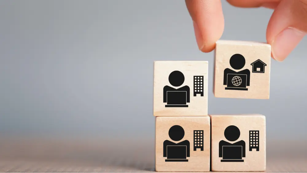 A hand building a stack of blocks that have icons representing a hybrid work team.