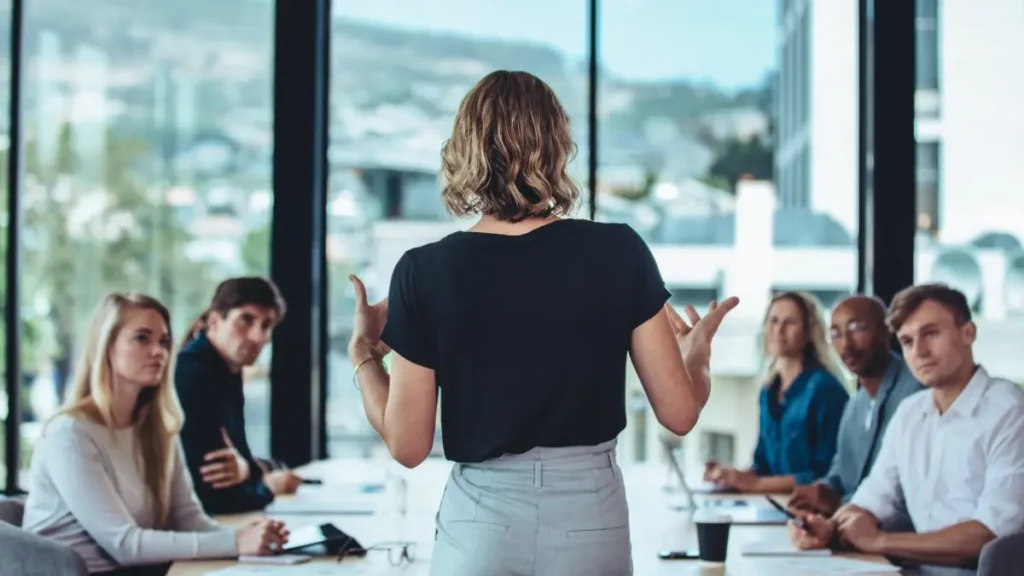 Businesswoman presenting to colleagues in a conference room with a city view in the background.
