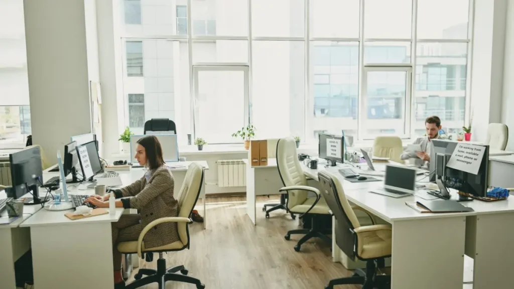 An office with two workers at their desks and several empty desks with notes saying the employees are working from home.