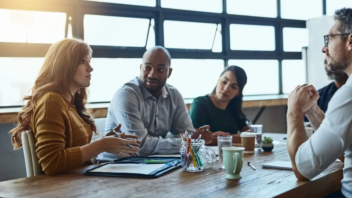 A diverse group of four professionals sits around a conference table in discussion. One woman is speaking while the others listen attentively, with notebooks, coffee cups, and pens on the table in a bright office setting.