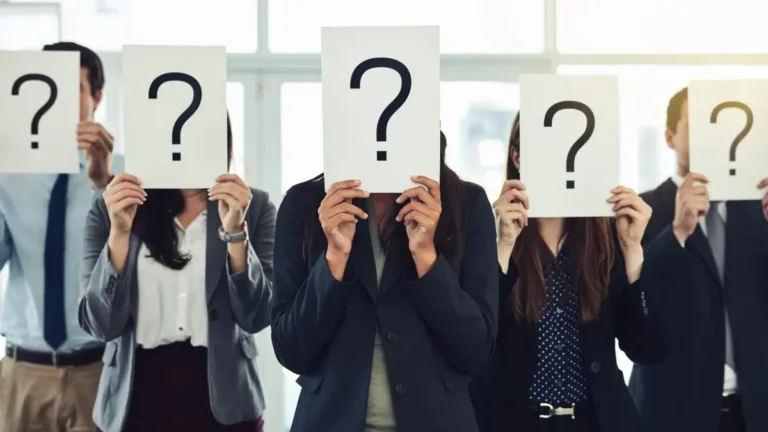 Group of business professionals holding up papers with large question marks covering their faces, symbolizing anonymity or uncertainty.
