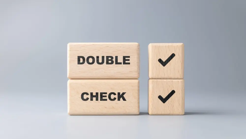 Two light wooden blocks stacked to read “DOUBLE CHECK,” next to two smaller wooden blocks with checkmark symbols, set against a neutral gray background.