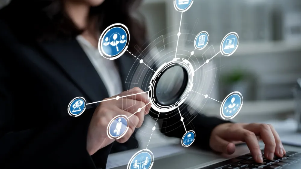 Person in business attire using a laptop while interacting with a large magnifying glass graphic overlaid on the screen. Around it are connected circular icons showing people, a résumé, a laptop, ratings stars, and meetings, suggesting recruitment, talent search, or workforce management.